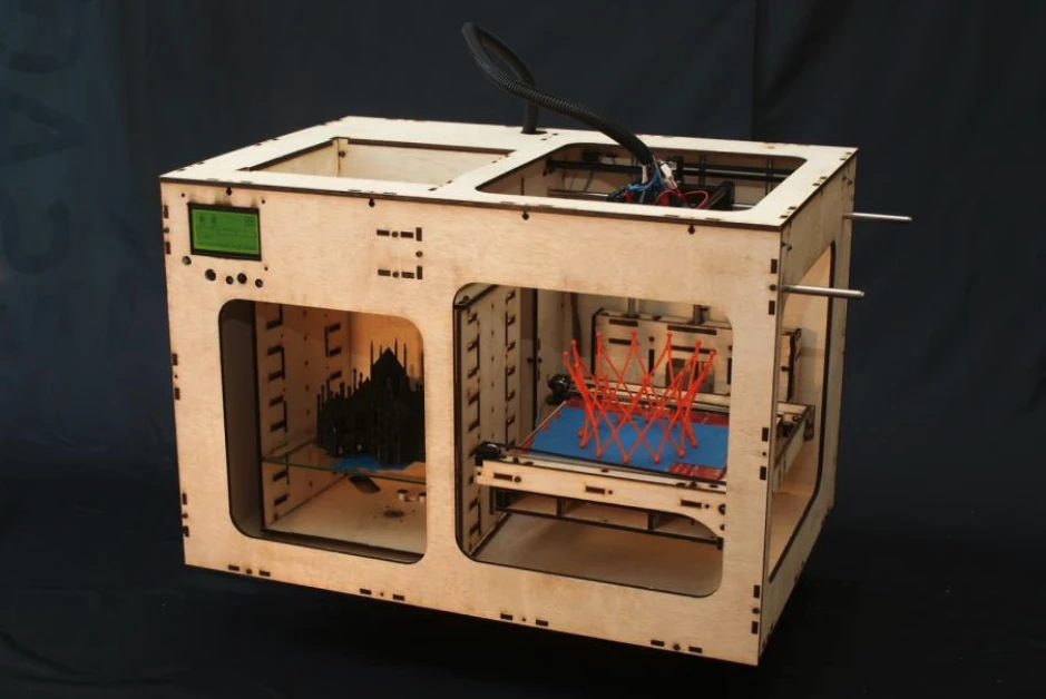 A laser-cut plywood 3D printer: a box with two windows in its front, a small green display at the top left, a black printed model in the left bay and a red lattice print on a blue bed in the right