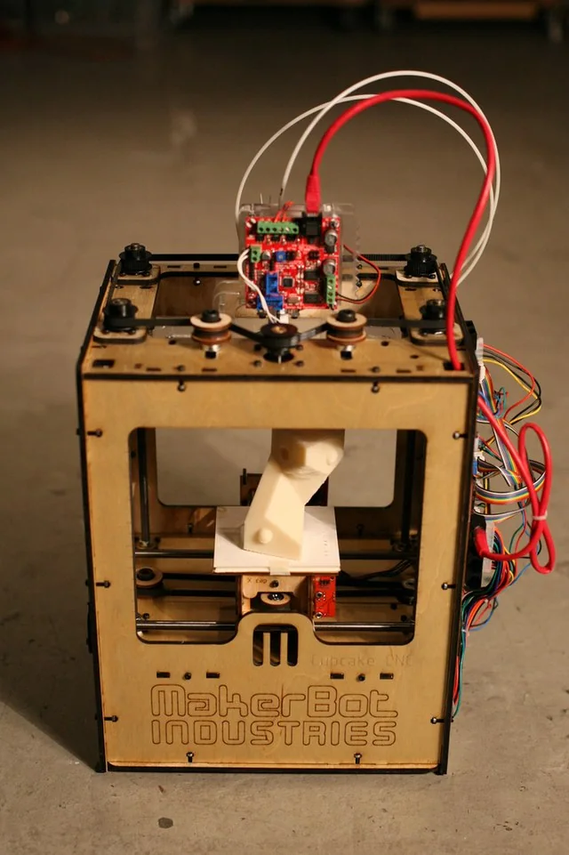A MakerBot Cupcake CNC: a small laser-cut plywood box with a red controller board and loose cables on top and a white print on its platform