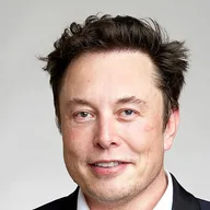 Elon Musk, head and shoulders, smiling slightly, in a dark jacket against a pale grey wall.