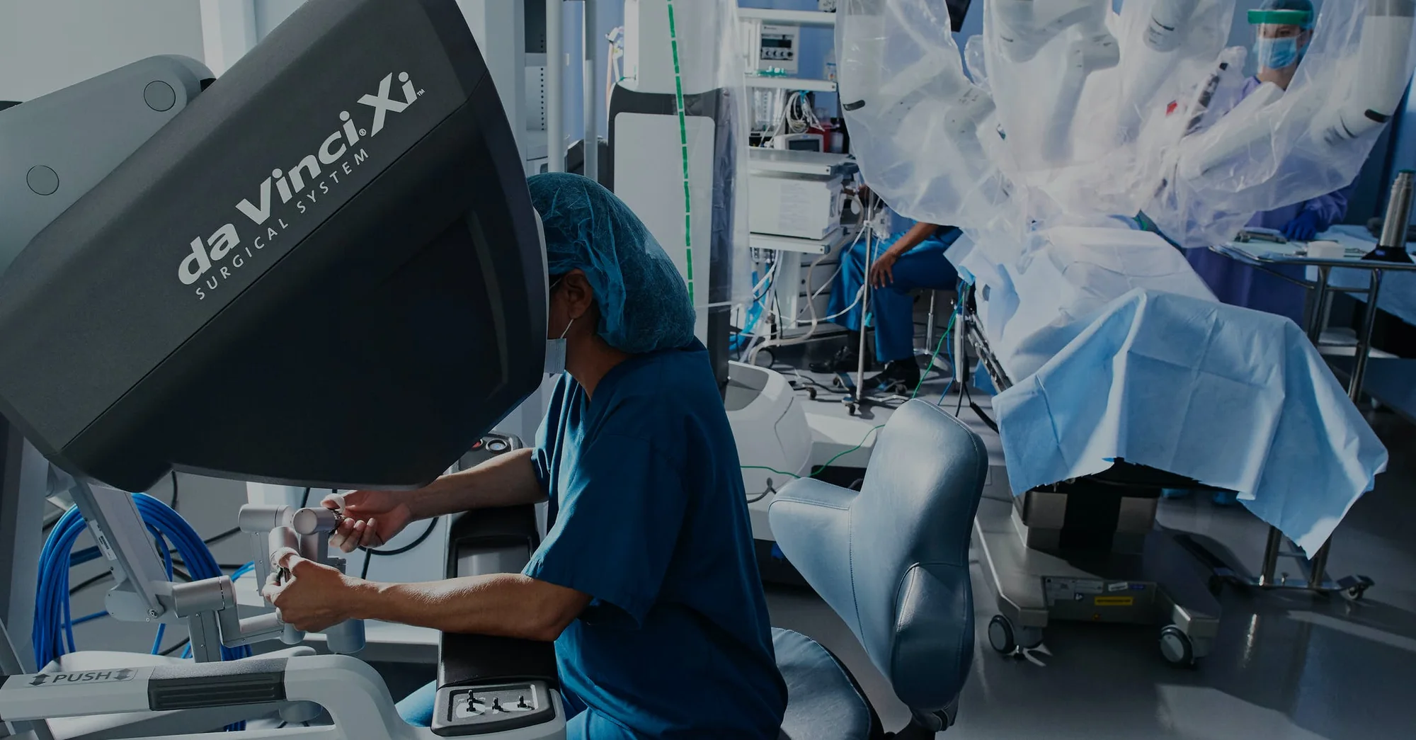 A surgeon in scrubs at the da Vinci Xi console, hands on its controls, with the draped robot arms over the patient behind
