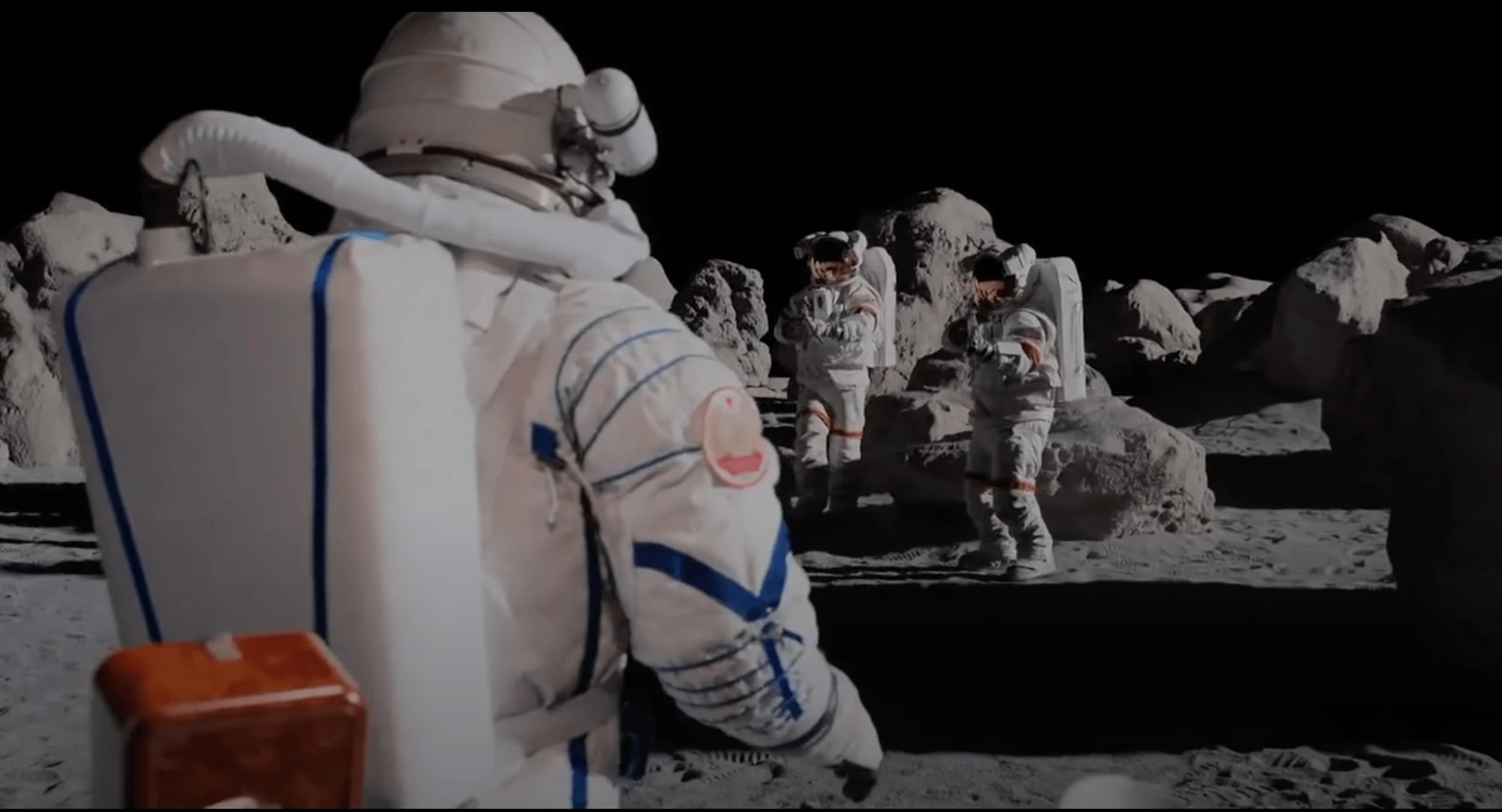 Over a Soviet cosmonaut's shoulder, two astronauts stand among boulders on the Moon
