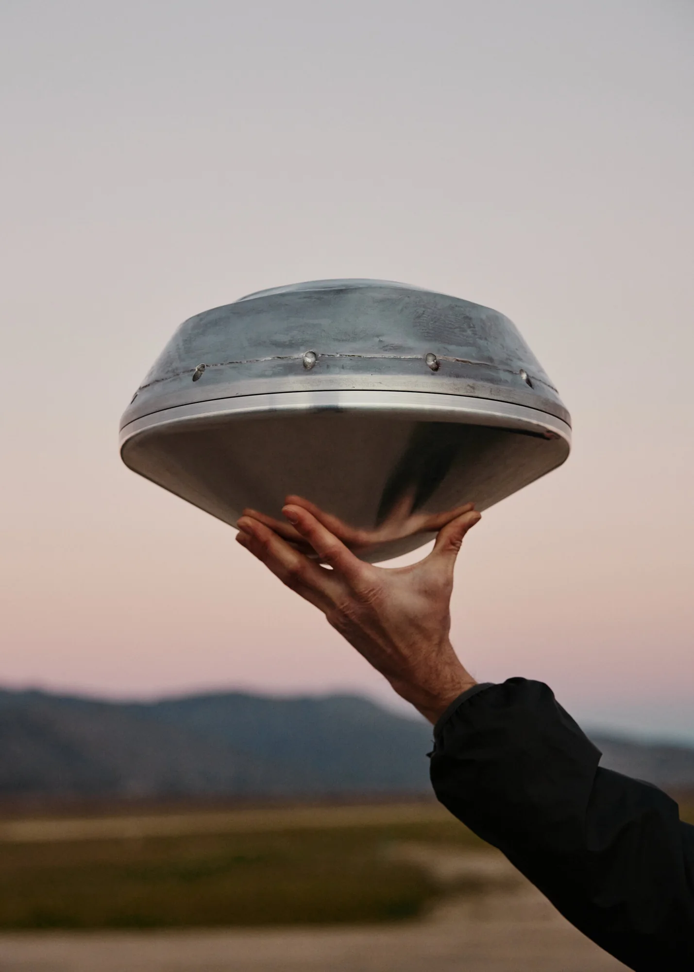 A hand holding up a small saucer-shaped metal capsule against a dusk sky