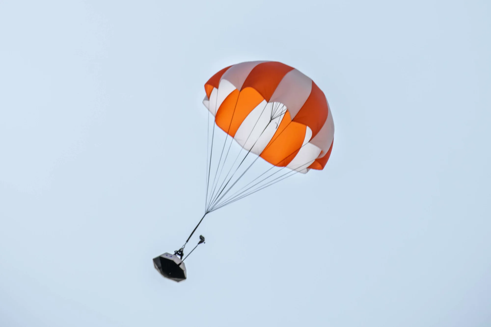 A small dark capsule hanging under an orange and white parachute against a pale sky